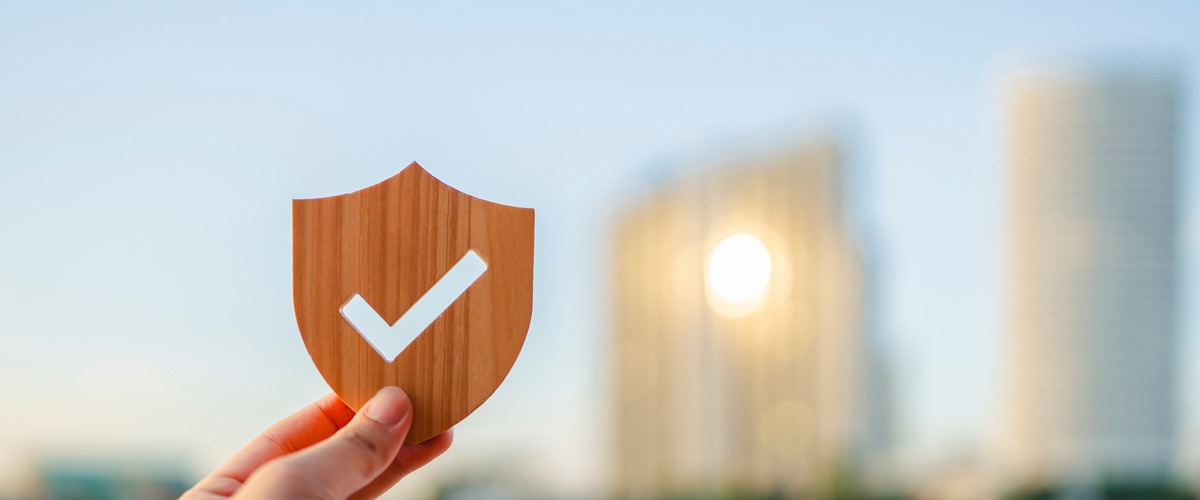 Bild zeigt Hand hält hölzernes Schild mit weißem Häkchen vor unscharfer Stadtkulisse verlinkt auf Blogbeitrag Risikolebensversicherung als Hinterbliebenenabsicherung